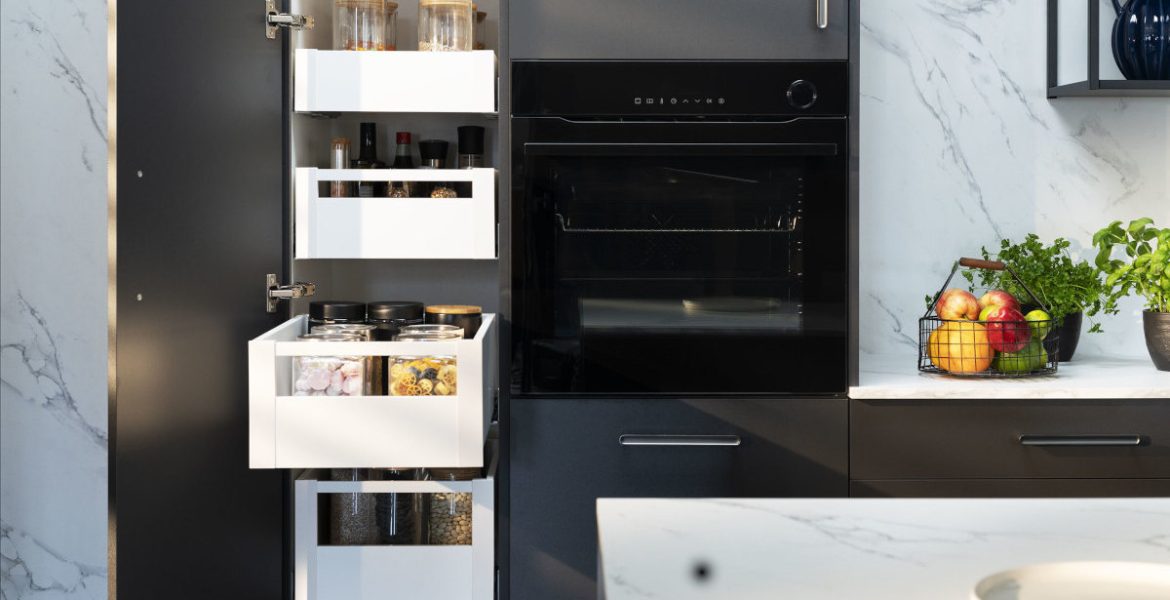 Wnętrze nowoczesnej, ciemnoszarej szafki kuchennej z kilkoma wysuniętymi białymi szufladami. W szufladach widać słoiki z sypkimi produktami, przyprawy, makaron i płatki. Po prawej stronie widać czarny piekarnik do zabudowy. Na blacie obok szafki stoi koszyk z owocami i zioła w doniczkach.