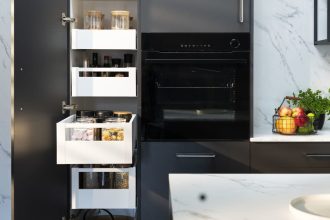 Wnętrze nowoczesnej, ciemnoszarej szafki kuchennej z kilkoma wysuniętymi białymi szufladami. W szufladach widać słoiki z sypkimi produktami, przyprawy, makaron i płatki. Po prawej stronie widać czarny piekarnik do zabudowy. Na blacie obok szafki stoi koszyk z owocami i zioła w doniczkach.