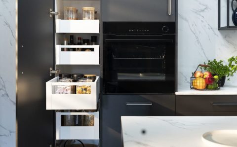 Wnętrze nowoczesnej, ciemnoszarej szafki kuchennej z kilkoma wysuniętymi białymi szufladami. W szufladach widać słoiki z sypkimi produktami, przyprawy, makaron i płatki. Po prawej stronie widać czarny piekarnik do zabudowy. Na blacie obok szafki stoi koszyk z owocami i zioła w doniczkach.