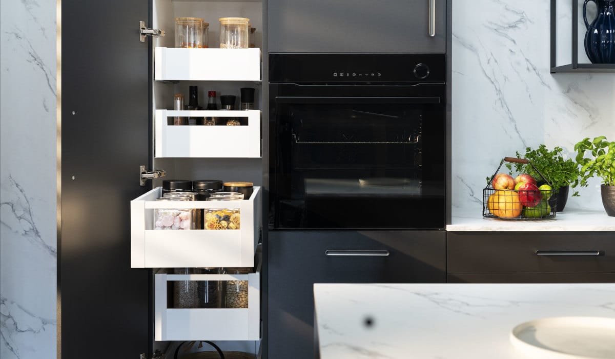 Wnętrze nowoczesnej, ciemnoszarej szafki kuchennej z kilkoma wysuniętymi białymi szufladami. W szufladach widać słoiki z sypkimi produktami, przyprawy, makaron i płatki. Po prawej stronie widać czarny piekarnik do zabudowy. Na blacie obok szafki stoi koszyk z owocami i zioła w doniczkach.