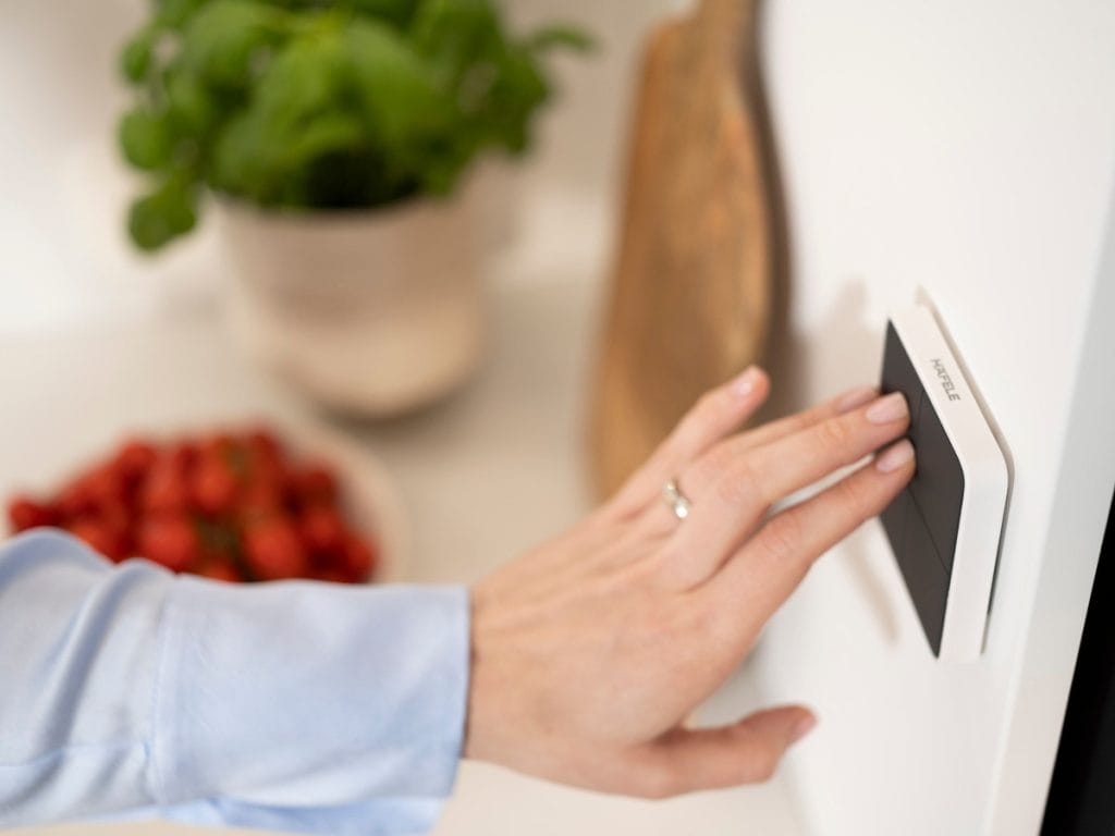 Zbliżenie dłoni kobiety w jasnoniebieskiej bluzce z pierścionkiem na palcu, naciskającej biały przełącznik z logo Häfele na białej ścianie. W rozmytym tle widać doniczkę z bazylią, drewnianą deskę i miskę truskawek.