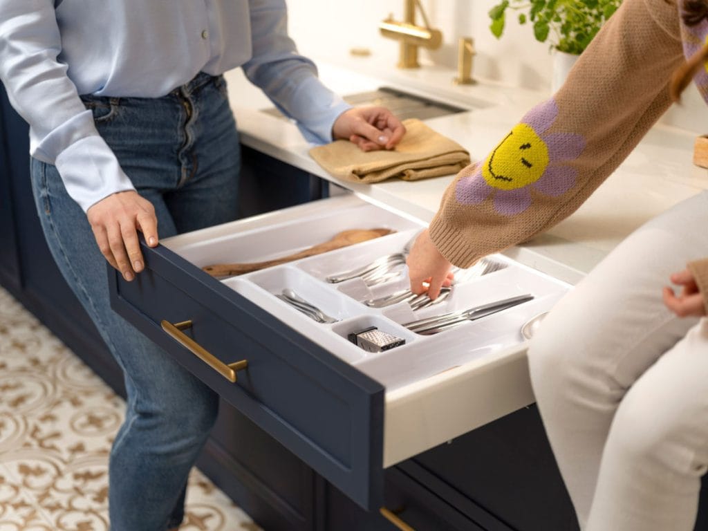 Zdjęcie dwóch osób stojących przy otwartej, ciemnoniebieskiej szufladzie kuchennej ze złotym uchwytem. Szuflada jest podświetlona i widać w niej biały wkład z ułożonymi sztućcami (widelcami). Jedna osoba (w niebieskiej koszuli) stoi obok szuflady, a druga (w beżowym swetrze z uśmiechem) sięga po widelec. W tle widać białą podłogę z brązowym, marokańskim wzorem.