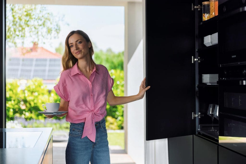 Kobieta o blond włosach ubrana w różową koszulę i dżinsy, trzymająca w jednej ręce filiżankę na spodeczku. Drugą ręką otwiera front czarnej, wysokiej szafki kuchennej w zabudowie, w której widać ułożone naczynia i szklanki. Tło zdjęcia stanowi duże okno wychodzące na zielony ogród.