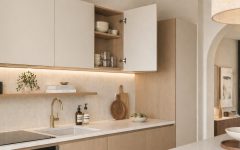 Szerokie ujęcie nowoczesnej, minimalistycznej kuchni w jasnych kolorach. Widać szafki wiszące z frontami w odcieniu złamanej bieli (jeden front jest otwarty, odsłaniając drewniane wnętrze z naczyniami i widocznymi zawiasami), dolną zabudowę w jasnym drewnie, biały blat, złoty kran, zlew, a na półce i blacie dekoracyjne akcesoria (butelki, deski do krojenia, miska z warzywami). W tle widać jadalnię z łukiem.