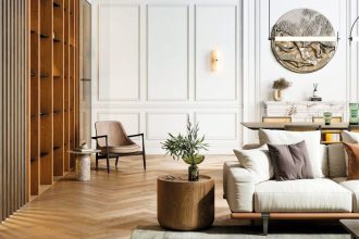 Wysoki salon z dużymi oknami, dzięki którym pomieszczenie zalane jest światłem, to doskonała sceneria dla aranżacji, w której drewno gra pierwsze skrzypce. Ozdobiona sztukateriami ściana, pomalowana odporną na plamy i nieestetyczne otarcia białą farbą Beckers Designer White, stanowi neutralne i niezwykle eleganckie tło dla przestrzeni, w której uwagę zwraca masywna drewniana zabudowa. Dębowa podłoga i drewniane meble uzupełniają look będący połączeniem przytulności i luksusu. Akcenty z kolorowego szkła i metalu wspierają płynne przejście między współczesnym designem, a statecznością drewnianych form. Źródło: Beckers.