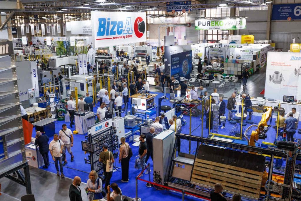 Wysokie, panoramiczne ujęcie tętniącej życiem hali targowej z perspektywy lotu ptaka. Przestrzeń jest wypełniona licznymi stoiskami wystawców, maszynami przemysłowymi i tłumem odwiedzających chodzących po niebieskiej wykładzinie. Centralnym punktem jest duże stoisko firmy BizeA z dużym podwieszanym banerem z logo.