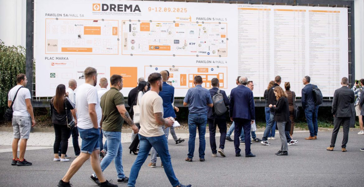 Ujęcie z poziomu gruntu grupy osób na zewnątrz, stojących przed dużą tablicą informacyjną targów DREMA 2025. Ludzie w swobodnym i biznesowym stroju idą lub stoją plecami do widza, patrząc na plan. Na tablicy widnieją napisy „DREMA 9-12.09.2025”, „PAWILON 5A/HALL 5A”, „PLAC MARKA” oraz mapa stoisk.