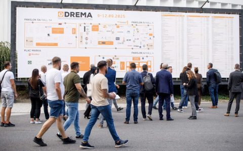 Ujęcie z poziomu gruntu grupy osób na zewnątrz, stojących przed dużą tablicą informacyjną targów DREMA 2025. Ludzie w swobodnym i biznesowym stroju idą lub stoją plecami do widza, patrząc na plan. Na tablicy widnieją napisy „DREMA 9-12.09.2025”, „PAWILON 5A/HALL 5A”, „PLAC MARKA” oraz mapa stoisk.