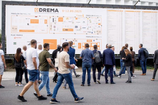 Ujęcie z poziomu gruntu grupy osób na zewnątrz, stojących przed dużą tablicą informacyjną targów DREMA 2025. Ludzie w swobodnym i biznesowym stroju idą lub stoją plecami do widza, patrząc na plan. Na tablicy widnieją napisy „DREMA 9-12.09.2025”, „PAWILON 5A/HALL 5A”, „PLAC MARKA” oraz mapa stoisk.