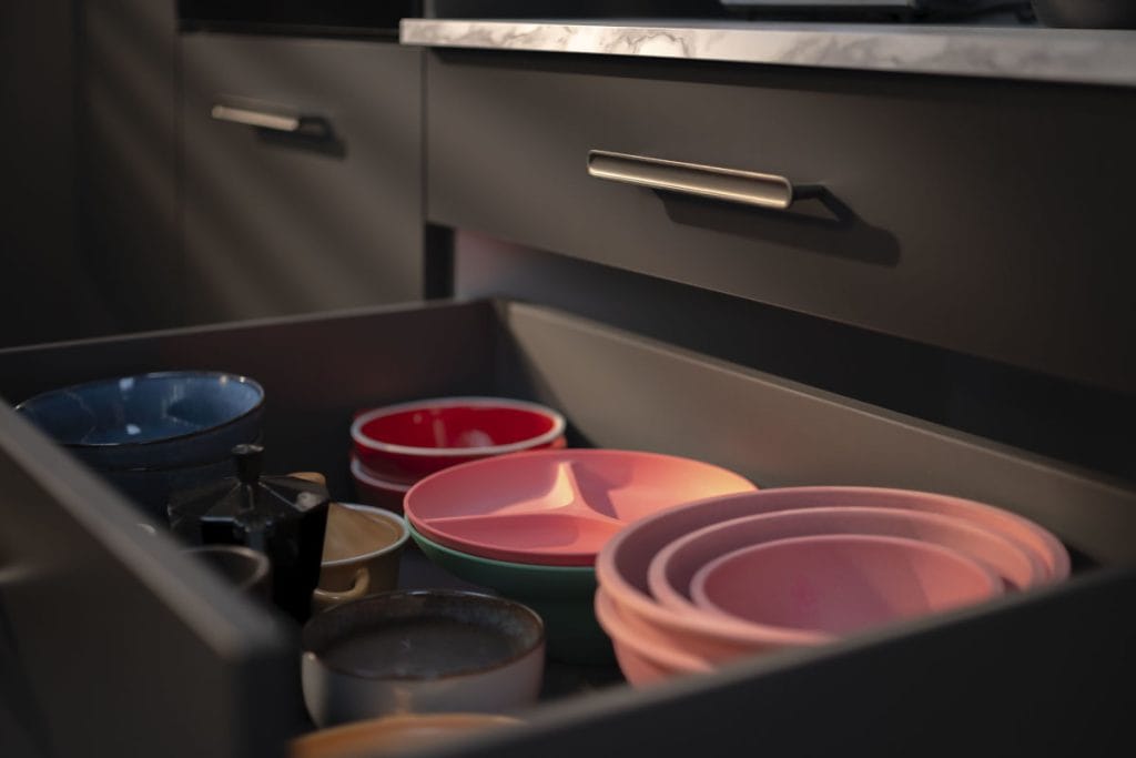 Zbliżenie na otwartą, antracytową szufladę kuchenną wypełnioną kolorową zastawą. Wewnątrz widoczne są starannie ułożone miski w kolorach niebieskim i czerwonym, różowe talerze z przegródkami oraz ceramiczne naczynia. Na zewnątrz widoczny jest minimalistyczny, podłużny uchwyt meblowy w kolorze ciemnego metalu oraz fragment białego, marmurowego blatu.