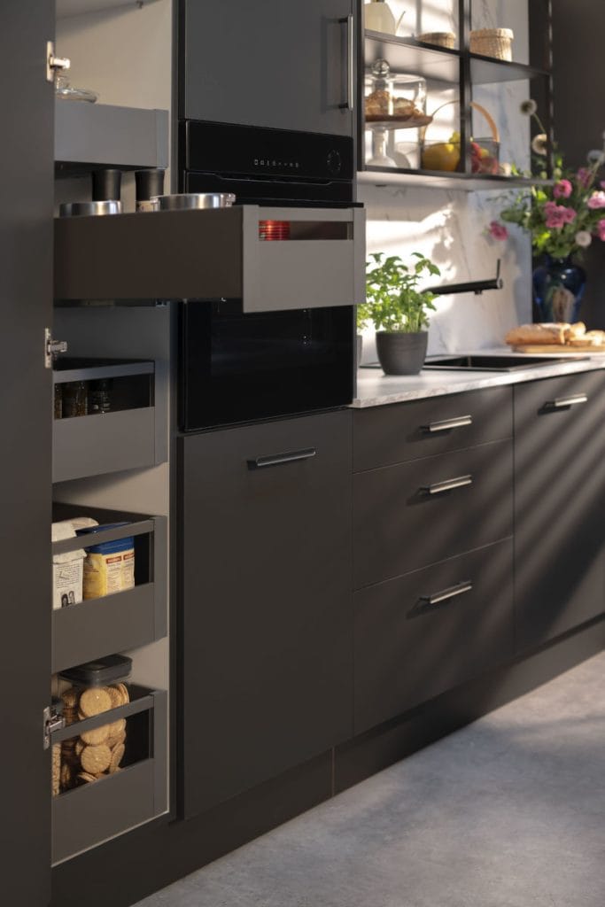 Wysoka zabudowa kuchenna w kolorze grafitowym z systemem wewnętrznych szuflad typu cargo. Cztery wysunięte szuflady prezentują przechowywanie produktów spożywczych: przypraw, płatków śniadaniowych oraz ciastek w słoikach. Obok znajduje się czarny piekarnik do zabudowy oraz fragment blatu z ziołami w doniczkach, zlewem i dekoracyjnym wazonem z kwiatami.
