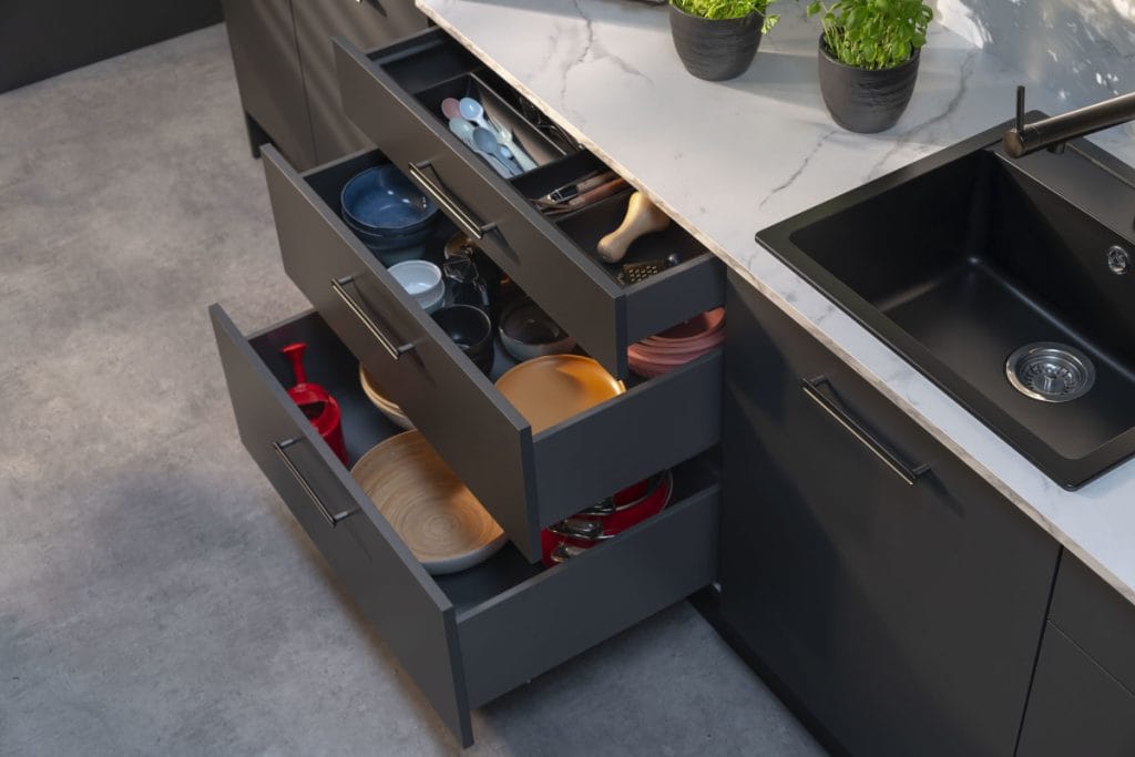 Widok z góry na otwartą sekcję trzech szuflad pod blatem kuchennym. Górna szuflada posiada organizer na sztućce, środkowa mieści kolorowe talerze i miski, a dolna, najgłębsza – duże misy i garnki. Obok znajduje się czarny zlew wpuszczany w jasny, żyłkowany blat oraz nowoczesna, czarna bateria kuchenna.