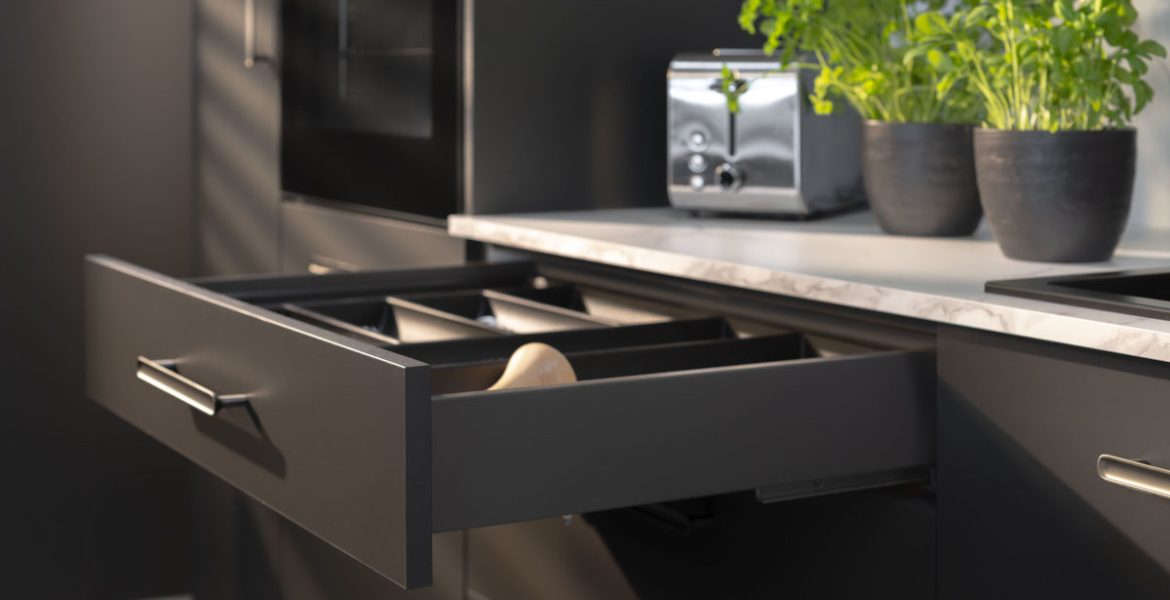 Detale nowoczesnej szuflady z systemem Matrix S w kolorze ciemnoszarym. Szuflada jest lekko wysunięta, prezentując czarny wkład organizacyjny. Na jasnym blacie w tle stoi chromowany toster oraz świeże zioła w ciemnych doniczkach. Światło padające z boku tworzy efektowne cienie na frontach mebli, podkreślając ich matowe wykończenie.