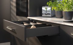 Detale nowoczesnej szuflady z systemem Matrix S w kolorze ciemnoszarym. Szuflada jest lekko wysunięta, prezentując czarny wkład organizacyjny. Na jasnym blacie w tle stoi chromowany toster oraz świeże zioła w ciemnych doniczkach. Światło padające z boku tworzy efektowne cienie na frontach mebli, podkreślając ich matowe wykończenie.
