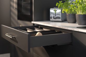 Detale nowoczesnej szuflady z systemem Matrix S w kolorze ciemnoszarym. Szuflada jest lekko wysunięta, prezentując czarny wkład organizacyjny. Na jasnym blacie w tle stoi chromowany toster oraz świeże zioła w ciemnych doniczkach. Światło padające z boku tworzy efektowne cienie na frontach mebli, podkreślając ich matowe wykończenie.