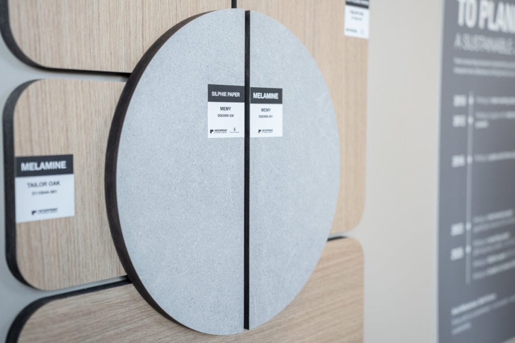 Zbliżenie na okrągłe i prostokątne próbki dekorów meblowych Interprint. Widoczne etykiety z nazwami technologii: „Silphie Paper”, „Melamine” oraz nazwami wzorów: „Memy” i „Tailor Oak”. Zdjęcie podkreśla teksturę materiałów i precyzję wykończenia.