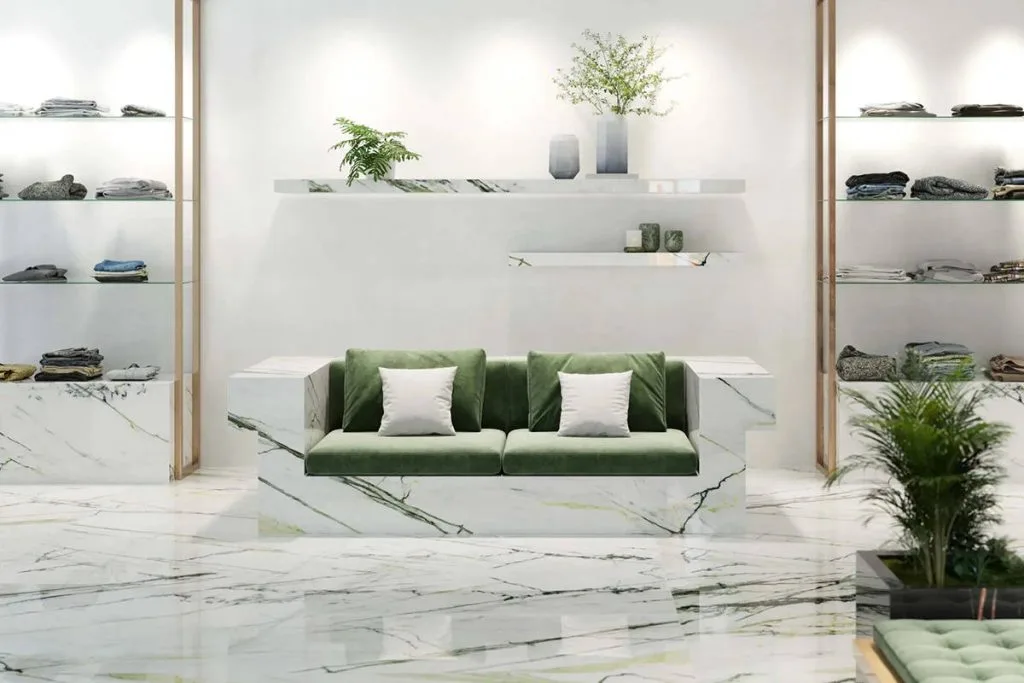 Eleganckie wnętrze butiku lub salonu ekspozycyjnego wykończone spiekiem kwarcowym Calacatta Green z charakterystycznym zielonym użyleniem. W centrum znajduje się geometryczna sofa obudowana tym samym materiałem, z zielonymi aksamitnymi siedziskami. W tle widoczne są białe ściany z minimalistycznymi półkami oraz regały z ekspozycją odzieży, co podkreśla luksusowy i nowoczesny charakter aranżacji.