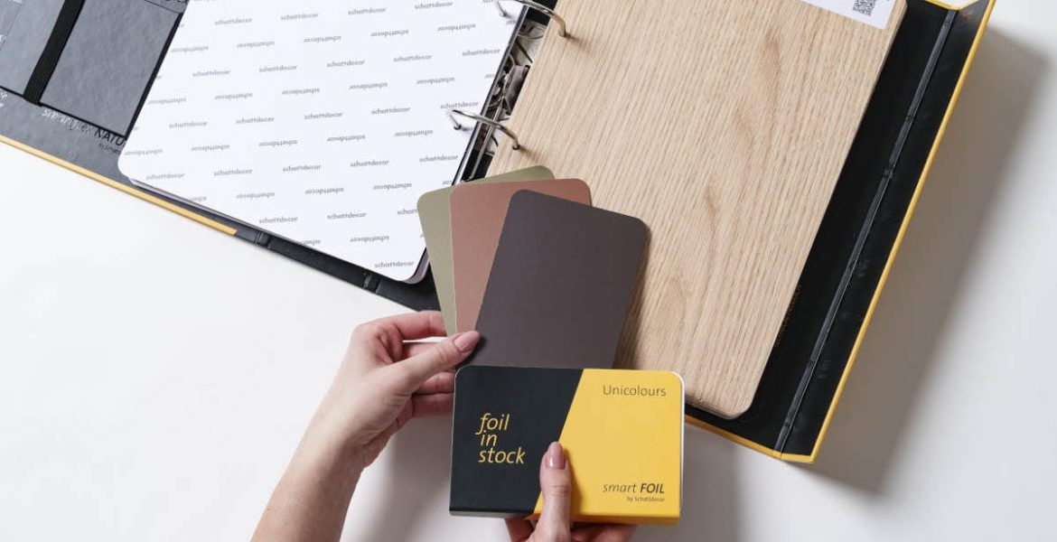 Otwarty wzornik produktów Schattdecor prezentujący folię Lindberg Oak oraz próbki kolorystyczne Unicolours z kolekcji smartfoil. Dłonie trzymające próbnik "foil in stock".