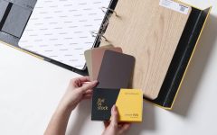 Otwarty wzornik produktów Schattdecor prezentujący folię Lindberg Oak oraz próbki kolorystyczne Unicolours z kolekcji smartfoil. Dłonie trzymające próbnik "foil in stock".