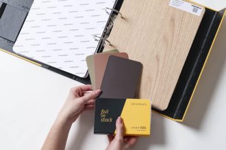 Otwarty wzornik produktów Schattdecor prezentujący folię Lindberg Oak oraz próbki kolorystyczne Unicolours z kolekcji smartfoil. Dłonie trzymające próbnik "foil in stock".