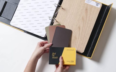 Otwarty wzornik produktów Schattdecor prezentujący folię Lindberg Oak oraz próbki kolorystyczne Unicolours z kolekcji smartfoil. Dłonie trzymające próbnik "foil in stock".