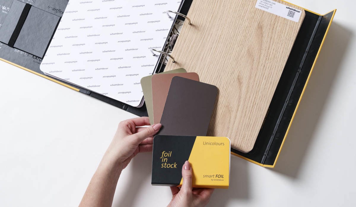 Otwarty wzornik produktów Schattdecor prezentujący folię Lindberg Oak oraz próbki kolorystyczne Unicolours z kolekcji smartfoil. Dłonie trzymające próbnik "foil in stock".