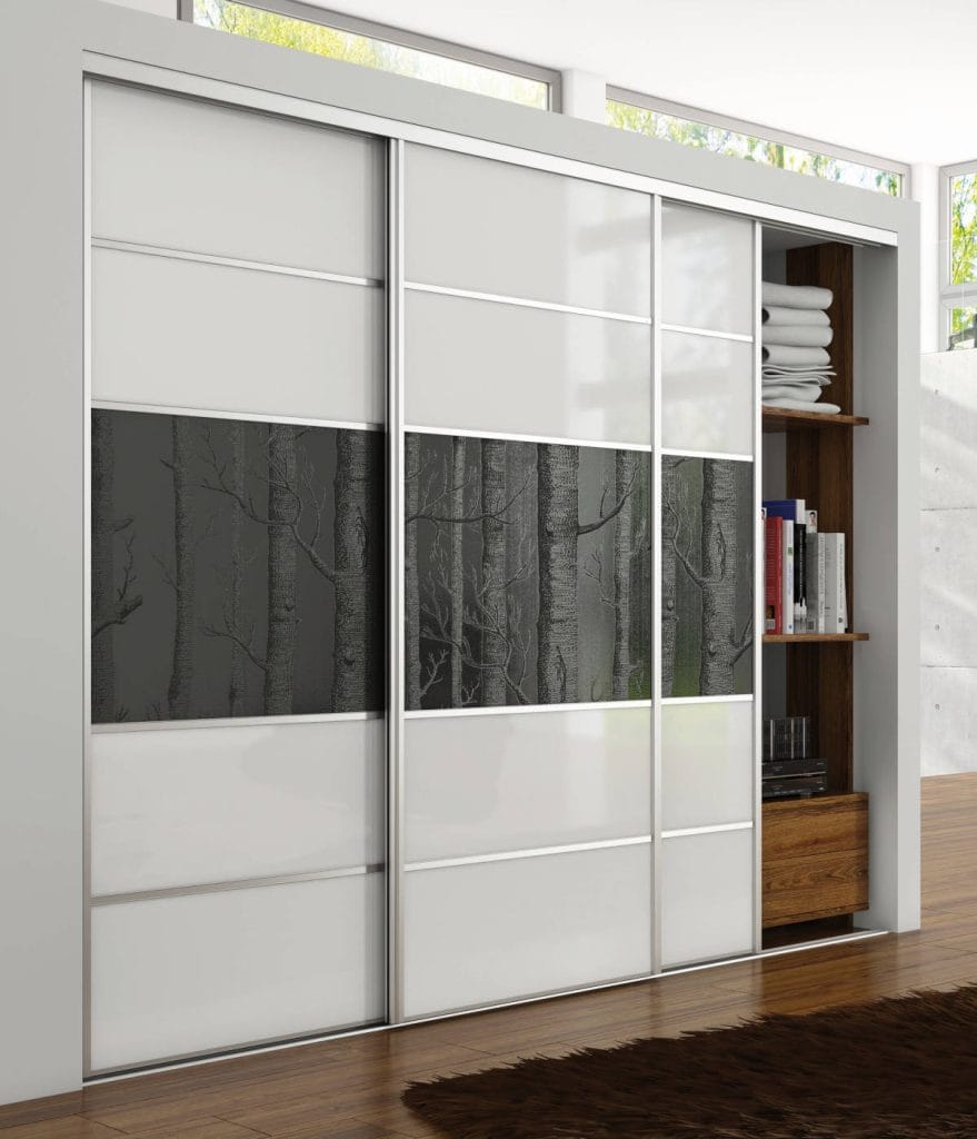 Trzyskrzydłowa szafa przesuwna w białej zabudowie. Fronty szafy wykonane z białego szkła przedzielonego poziomymi profilami, z centralnym pasem dekoracyjnym przedstawiającym grafikę czarno-białego lasu. Jedno skrzydło odsłania wnętrze z drewnianymi półkami.