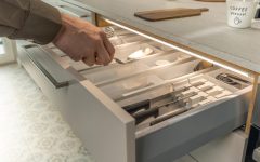 Zbliżenie na otwartą szufladę kuchenną z wkładem na sztućce, oświetloną systemem Häfele Loox5. Dłoń wyjmuje widelec z przegrody. Oświetlenie LED zamontowane pod krawędzią blatu roboczego.