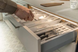 Zbliżenie na otwartą szufladę kuchenną z wkładem na sztućce, oświetloną systemem Häfele Loox5. Dłoń wyjmuje widelec z przegrody. Oświetlenie LED zamontowane pod krawędzią blatu roboczego.