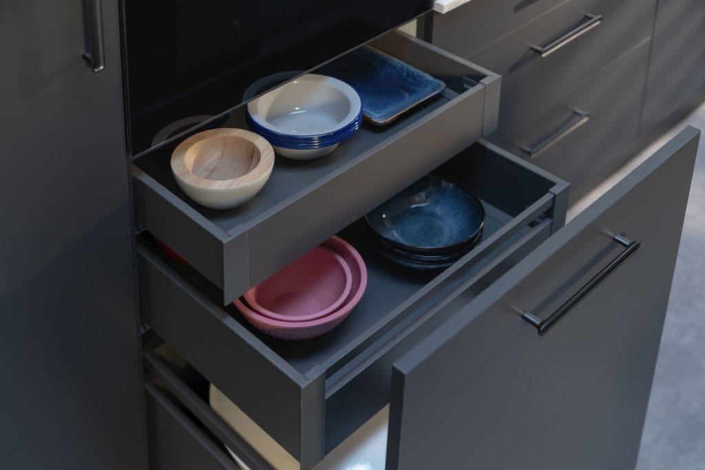 Ciemnoszare szuflady wewnętrzne Häfele Matrix S Slim wysunięte z wysokiej zabudowy kuchennej. W szufladach uporządkowane kolorowe miski ceramiczne i drewniane. Minimalistyczny design z cienkimi ściankami bocznymi.