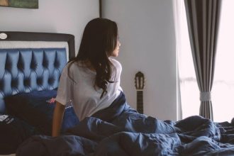 Kobieta siedząca na komfortowym łóżku w jasnej sypialni patrząca przez okno. Łóżka sypialniane dopasowane do Twoich potrzeb – wybierz model z tapicerowanym zagłówkiem, aby zadbać o komfortowy i zdrowy sen.
