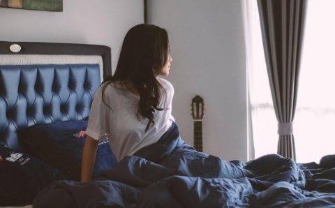 Kobieta siedząca na komfortowym łóżku w jasnej sypialni patrząca przez okno. Łóżka sypialniane dopasowane do Twoich potrzeb – wybierz model z tapicerowanym zagłówkiem, aby zadbać o komfortowy i zdrowy sen.