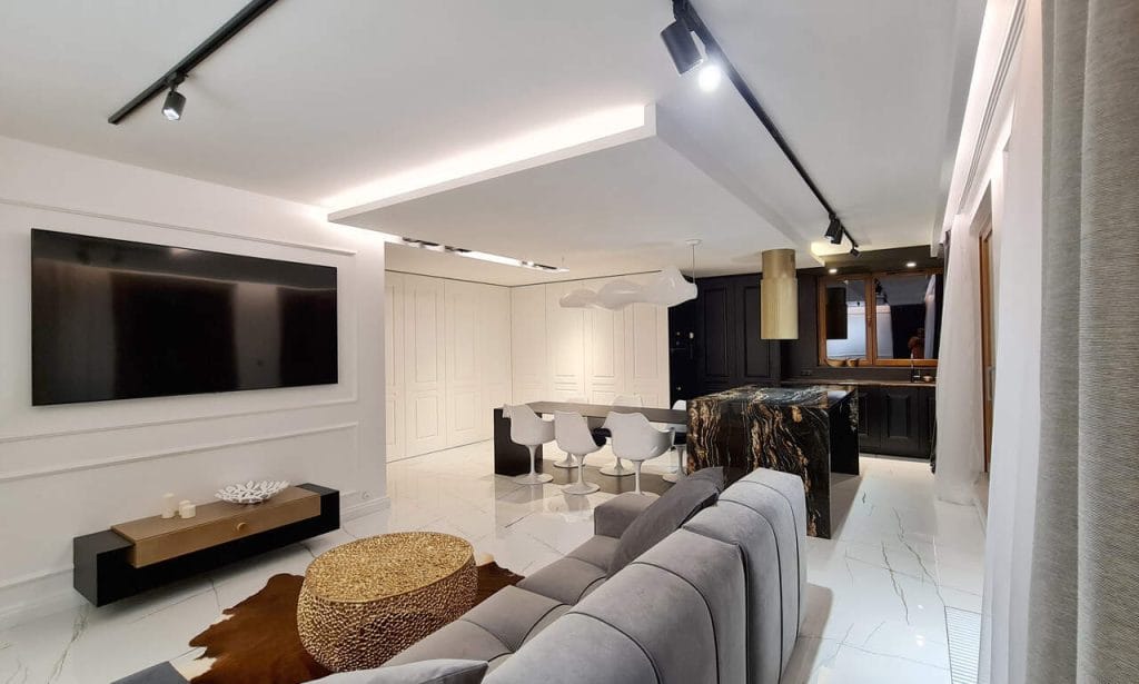 Luksusowy apartament dwupoziomowy o powierzchni ok. 140 m2 przeznaczony pod wynajem dla klienta premium, zlokalizowany w prestiżowej enklawie Magnoliowe Ogrody na Woli Justowskiej w Krakowie. W tej inspirowanej stylem Glamour realizacji dominuje biel skontrastowana sporą dawką czerni i ocieplona akcentami brązów i złota. Klasycznie frezowane białe fronty dopasowano do wzoru sztukaterii zastosowanych na ścianach. Za nimi kryją się nie tylko szafy i półki, ale też dyskretne przejścia do gabinetu, WC i schowka gospodarczego. Monolityczną, kamienną bryłę wyspy kuchennej przebija cienka jak żyletka płaszczyzna stołu  wykonanego z utwardzanego, lakierowanego aluminium. Możliwe jest wysunięcie go ze szczeliny w wyspie i uzyskanie dodatkowego miejsca dla łącznie 10 osób. Projekt i realizacja: Swałtek Design Studio.