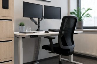 Nowoczesne stanowisko pracy z ergonomicznym biurkiem o regulowanej wysokości, dwoma monitorami na ramieniu, czarnym krzesłem siatkowym i szafką na dokumenty z wbudowanym tabletem.