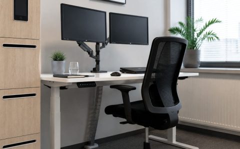 Nowoczesne stanowisko pracy z ergonomicznym biurkiem o regulowanej wysokości, dwoma monitorami na ramieniu, czarnym krzesłem siatkowym i szafką na dokumenty z wbudowanym tabletem.