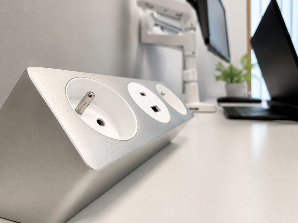 Zbliżenie na srebrny, skośny panel na biurku, zawierający jedno gniazdo zasilania Schuko oraz dwa porty ładowania USB (Typ-A i Typ-C). W tle widać monitory i laptopa.