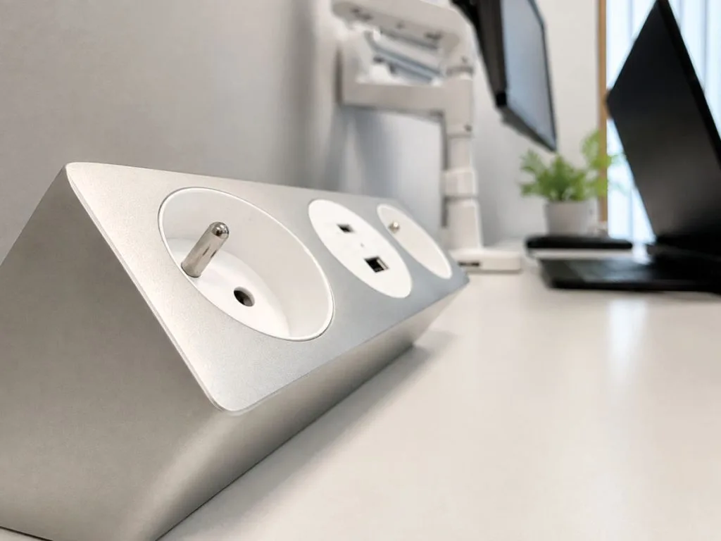 Zbliżenie na srebrny, skośny panel na biurku, zawierający jedno gniazdo zasilania Schuko oraz dwa porty ładowania USB (Typ-A i Typ-C). W tle widać monitory i laptopa.