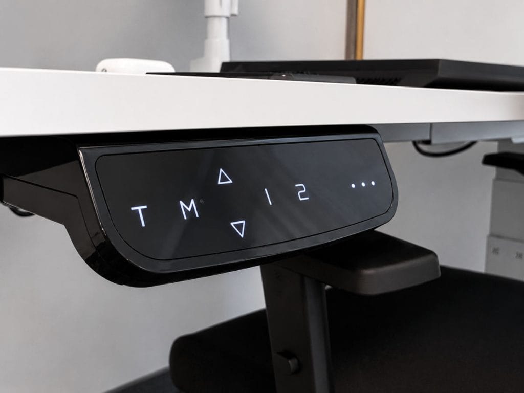 Zbliżenie na czarny, cyfrowy panel sterowania zamontowany pod blatem białego biurka elektrycznego. Panel wyświetla podświetlane symbole: strzałki góry/dół, litery 'T' i 'M', cyfry '1' i '2' oraz kropki.