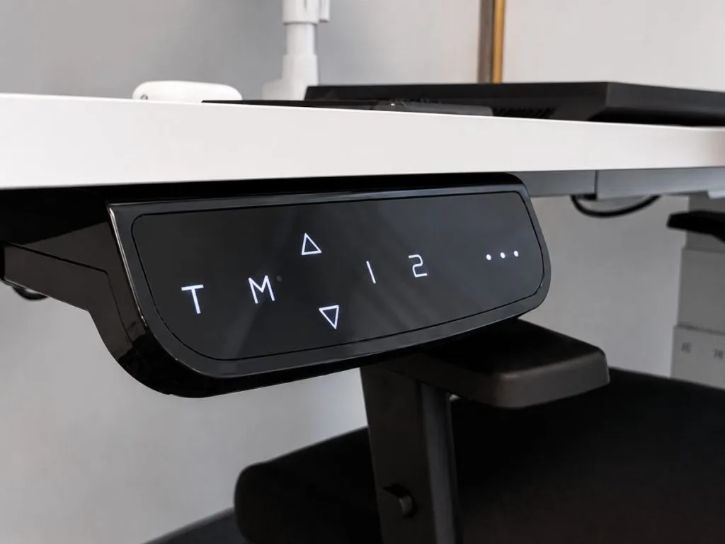 Zbliżenie na czarny, cyfrowy panel sterowania zamontowany pod blatem białego biurka elektrycznego. Panel wyświetla podświetlane symbole: strzałki góry/dół, litery 'T' i 'M', cyfry '1' i '2' oraz kropki.
