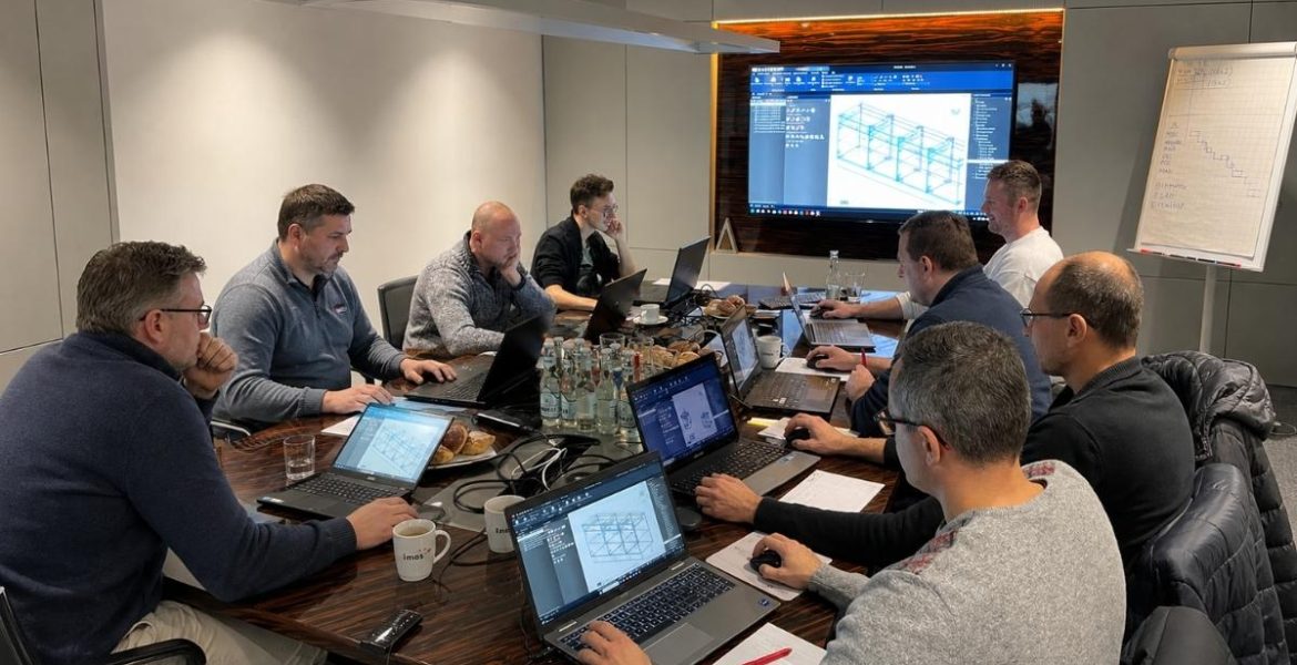 Zdjęcie spotkania biznesowego, na którym zespół ekspertów analizuje modele 3D i dane techniczne na ekranach laptopów i monitorze ściennym w ramach projektu wdrożenia systemu Imos.