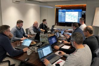 Zdjęcie spotkania biznesowego, na którym zespół ekspertów analizuje modele 3D i dane techniczne na ekranach laptopów i monitorze ściennym w ramach projektu wdrożenia systemu Imos.