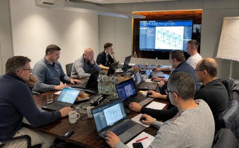 Zdjęcie spotkania biznesowego, na którym zespół ekspertów analizuje modele 3D i dane techniczne na ekranach laptopów i monitorze ściennym w ramach projektu wdrożenia systemu Imos.