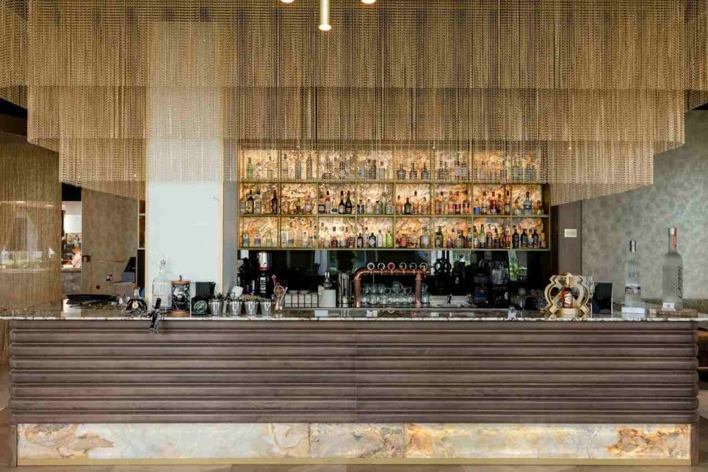 Strefa barowa we wnętrzu restauracji Salvia, zaprojektowanym przez pracownię Tremend, przyciąga wzrok monumentalną, kaskadową instalacją ze złotych łańcuchów oraz podświetlanym frontem lady o głębokiej, geometrycznej strukturze drewna. To kwintesencja nowoczesnego luksusu w segmencie HoReCa. Fot. Bartek Barczyk, materiały prasowe Tremend.