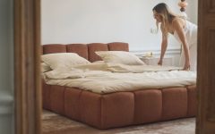 Kobieta ścieli nowoczesne łóżko tapicerowane w kolorze rdzawego brązu, od polskiej marki wnętrzarskiej Selsey.