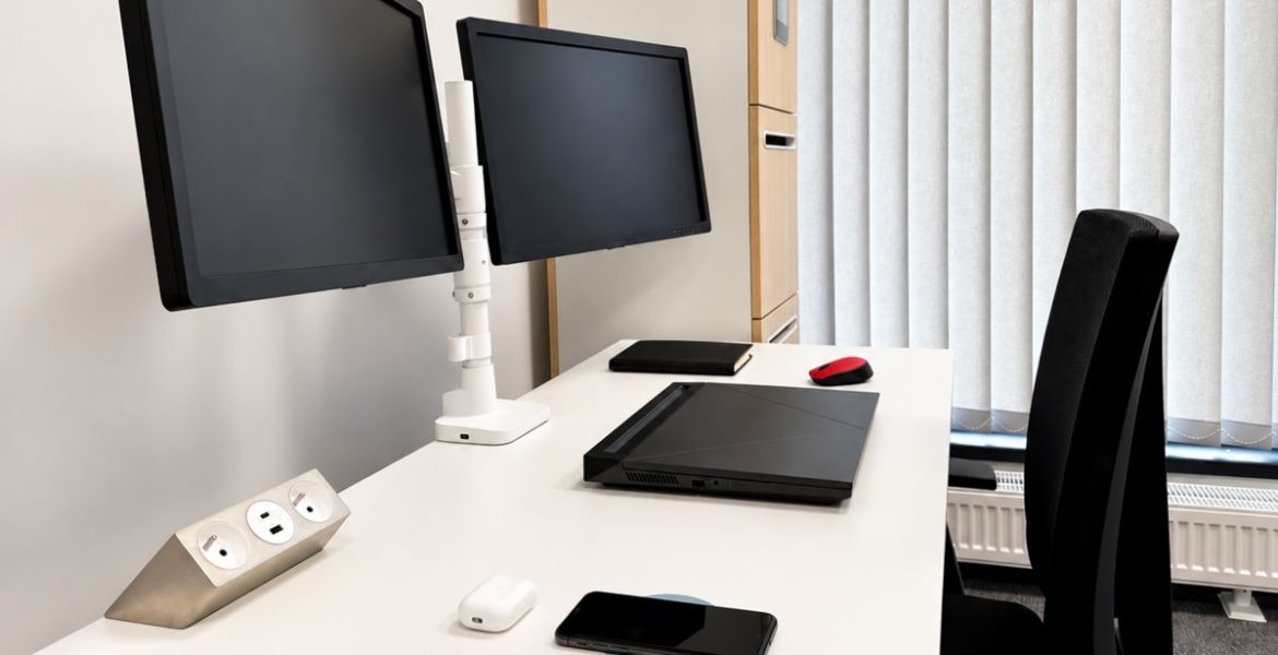 Modernistyczne stanowisko pracy z białym biurkiem, dwoma monitorami, laptopem oraz natynkowym gniazdem meblowym z portami USB.
