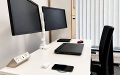 Modernistyczne stanowisko pracy z białym biurkiem, dwoma monitorami, laptopem oraz natynkowym gniazdem meblowym z portami USB.