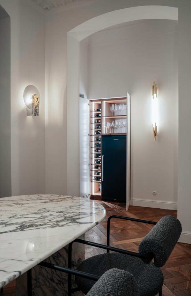 idok z jadalni na wbudowany bar w apartamencie z klasycznymi detalami. Na pierwszym planie stoi stół z marmuru Arabescato i krzesło z szarej tkaniny boucle. Przez łukowate przejście widać podświetloną szafkę barową z winem, kieliszkami do wina i chłodziarką na napoje. Po obu stronach przejścia znajdują się designerskie kinkiety, a na podłodze parkiet.